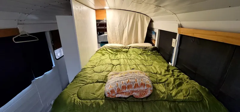Bed inside the Camp Calypso skoolie with a green comforter under a white plank ceiling