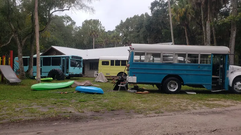 Kayaks on the grass beside a blue skoolie, a yellow bus and a turquoise transit bus